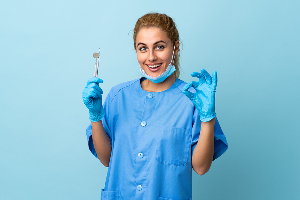 Young woman dentist holding tools over isolated blue background showing an ok sign with fingers dentifree-neocamino